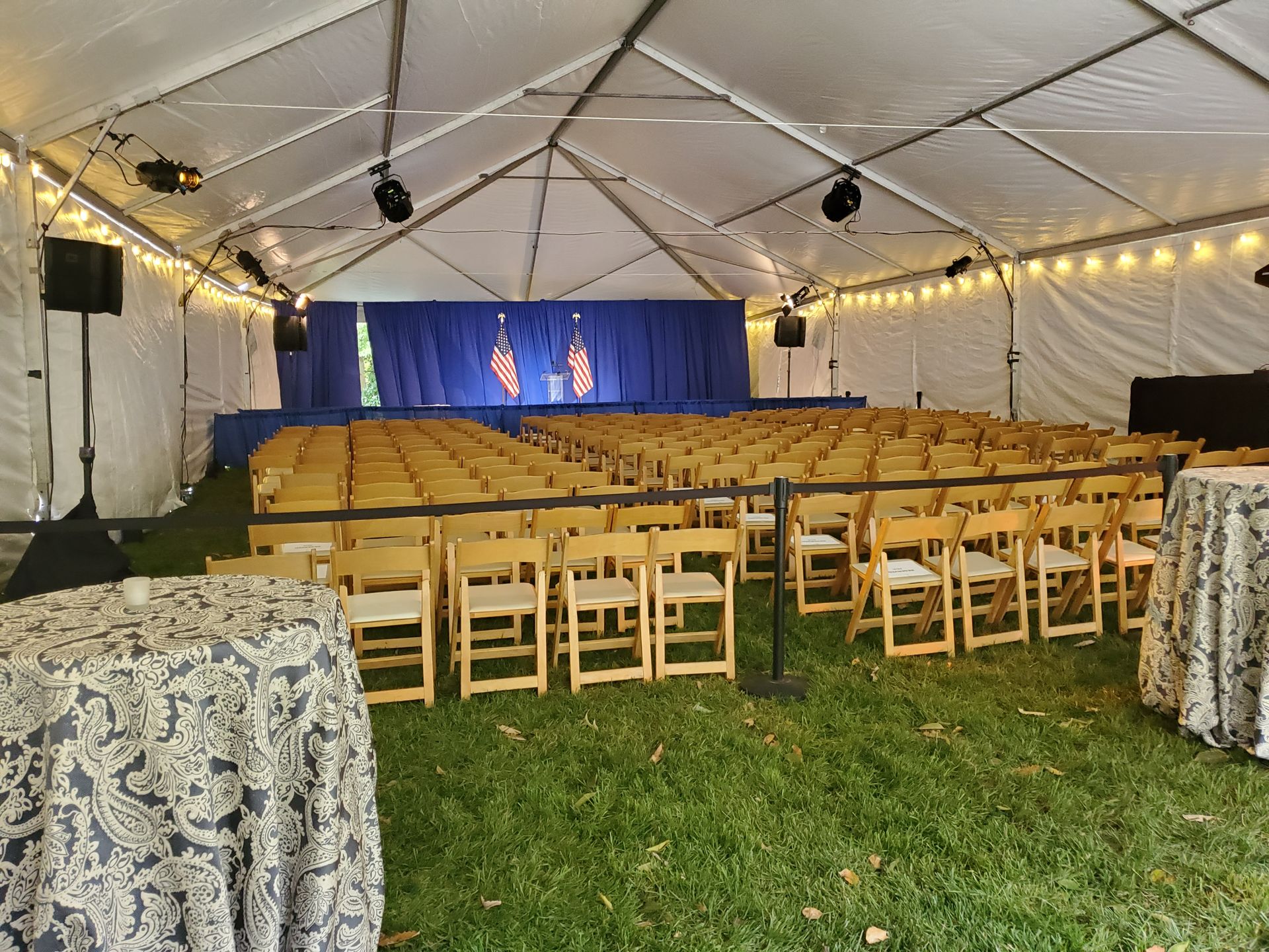 Inside a white tent: Rows of chairs face a stage with a blue backdrop. Two tables with patterned tablecloths.