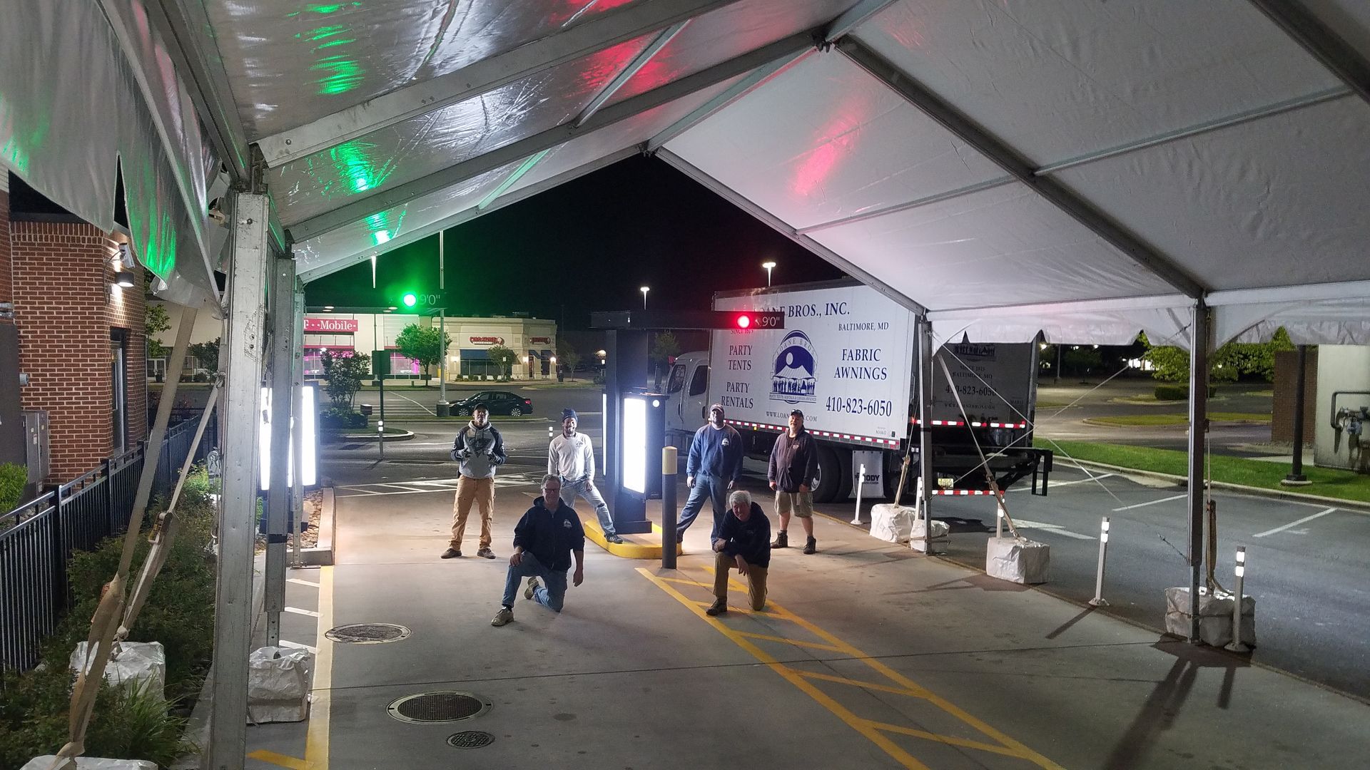 People in dark clothing gather under a tent, near a truck with emergency lights. Nighttime, exterior.
