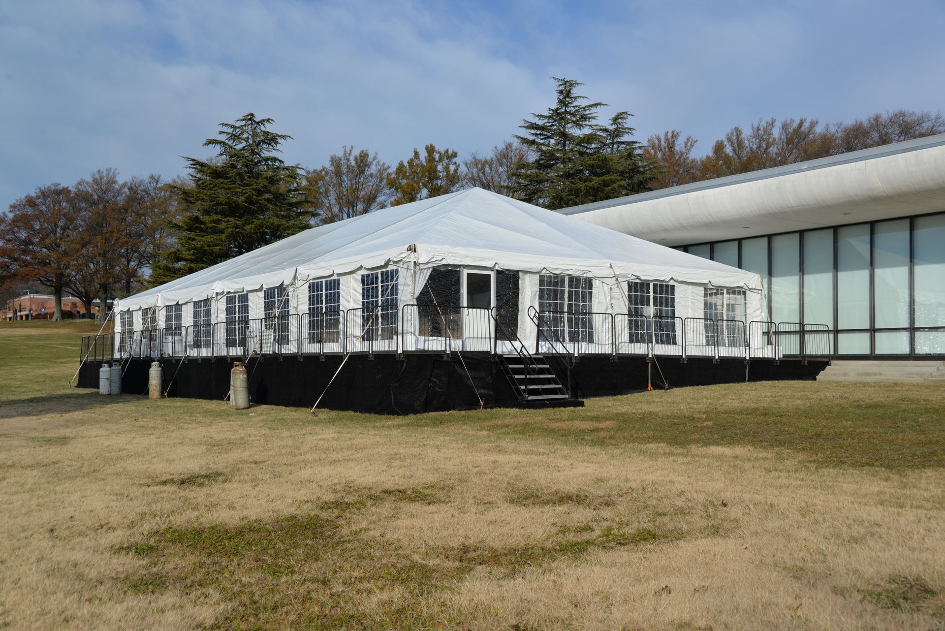 White tent on black platform, next to a modern building, on brown grass.