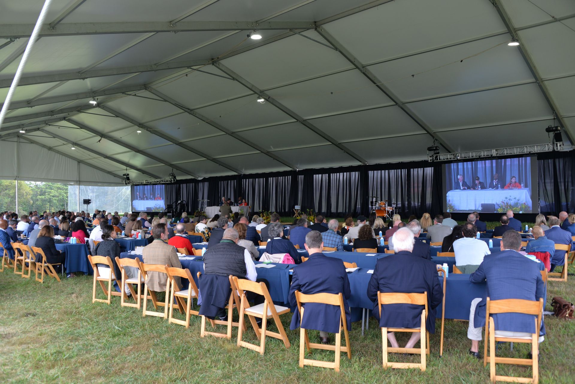 Large outdoor event tent with people seated at tables, watching a presentation on screens.