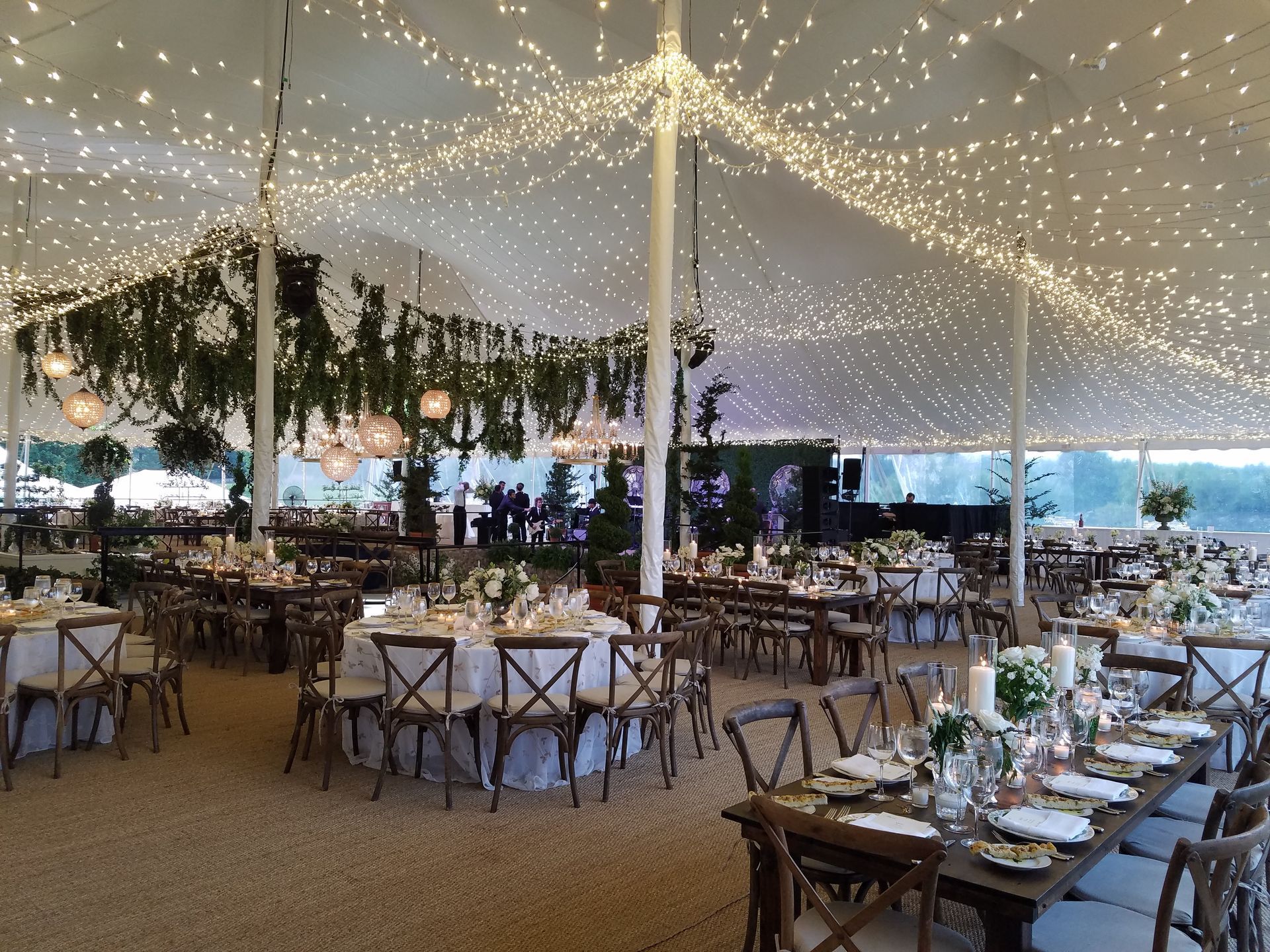 Wedding reception under a tent with string lights, tables set with floral centerpieces, and chairs.