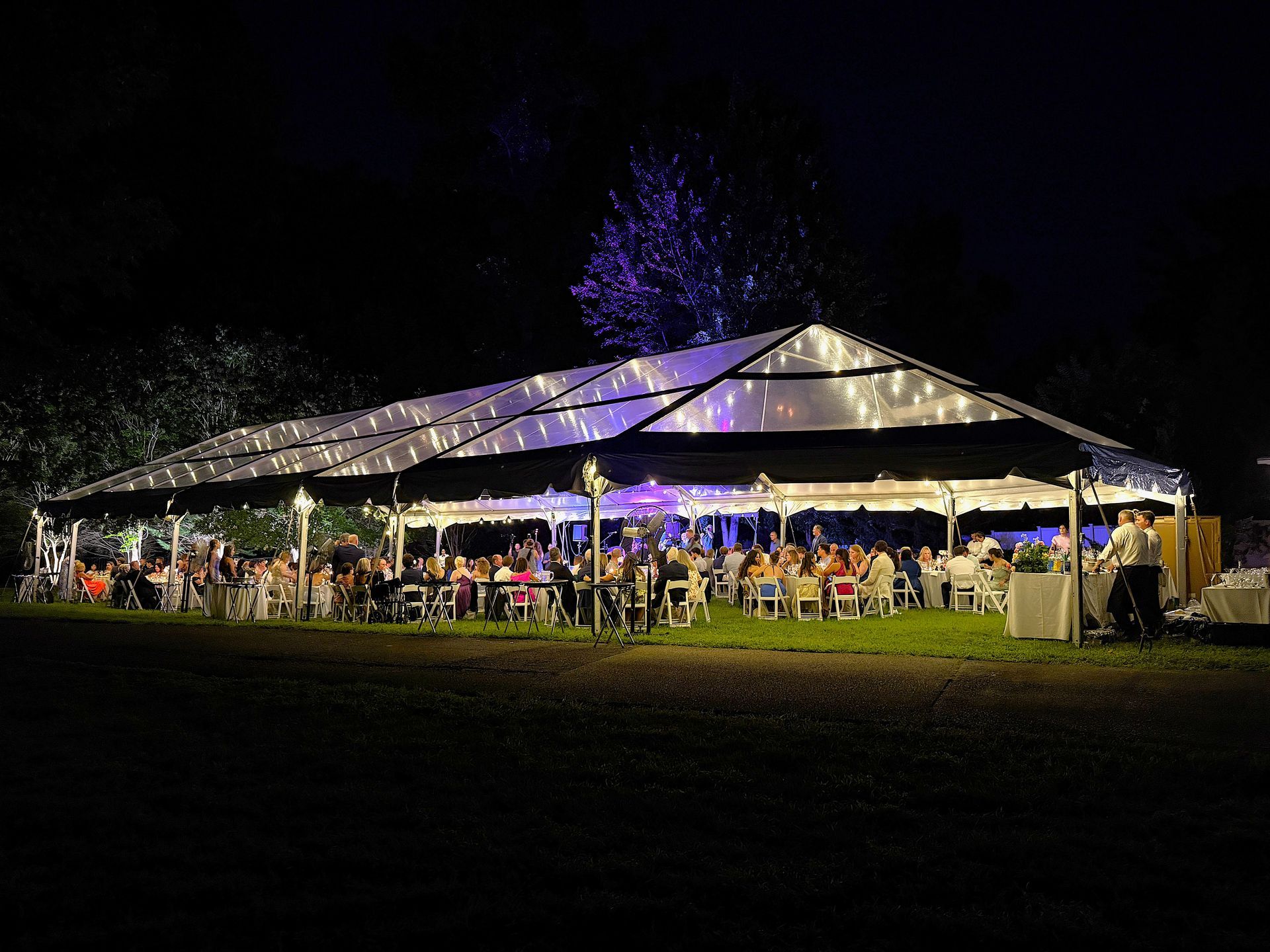 Large outdoor party under a tent at night, illuminated by string lights; guests are seated at tables.