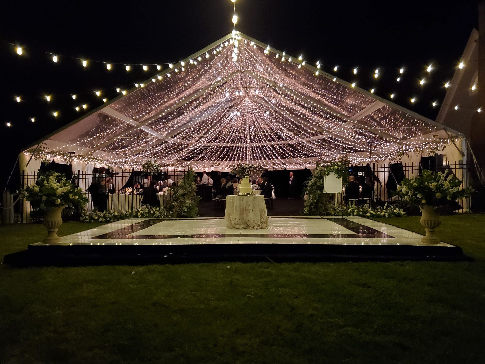 A clear tent lit with fairy lights at night, set up for an event. A cake sits in the center, surrounded by greenery.