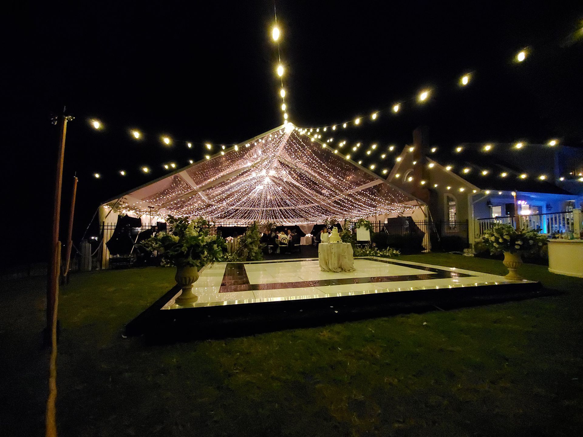 A nighttime outdoor wedding reception under a clear tent with string lights, a dance floor, and flowers.