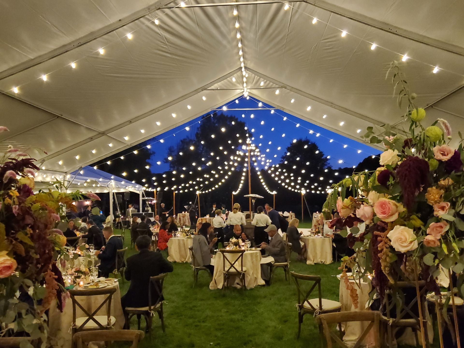 Wedding reception under a tent with string lights, guests seated at tables, and floral arrangements.