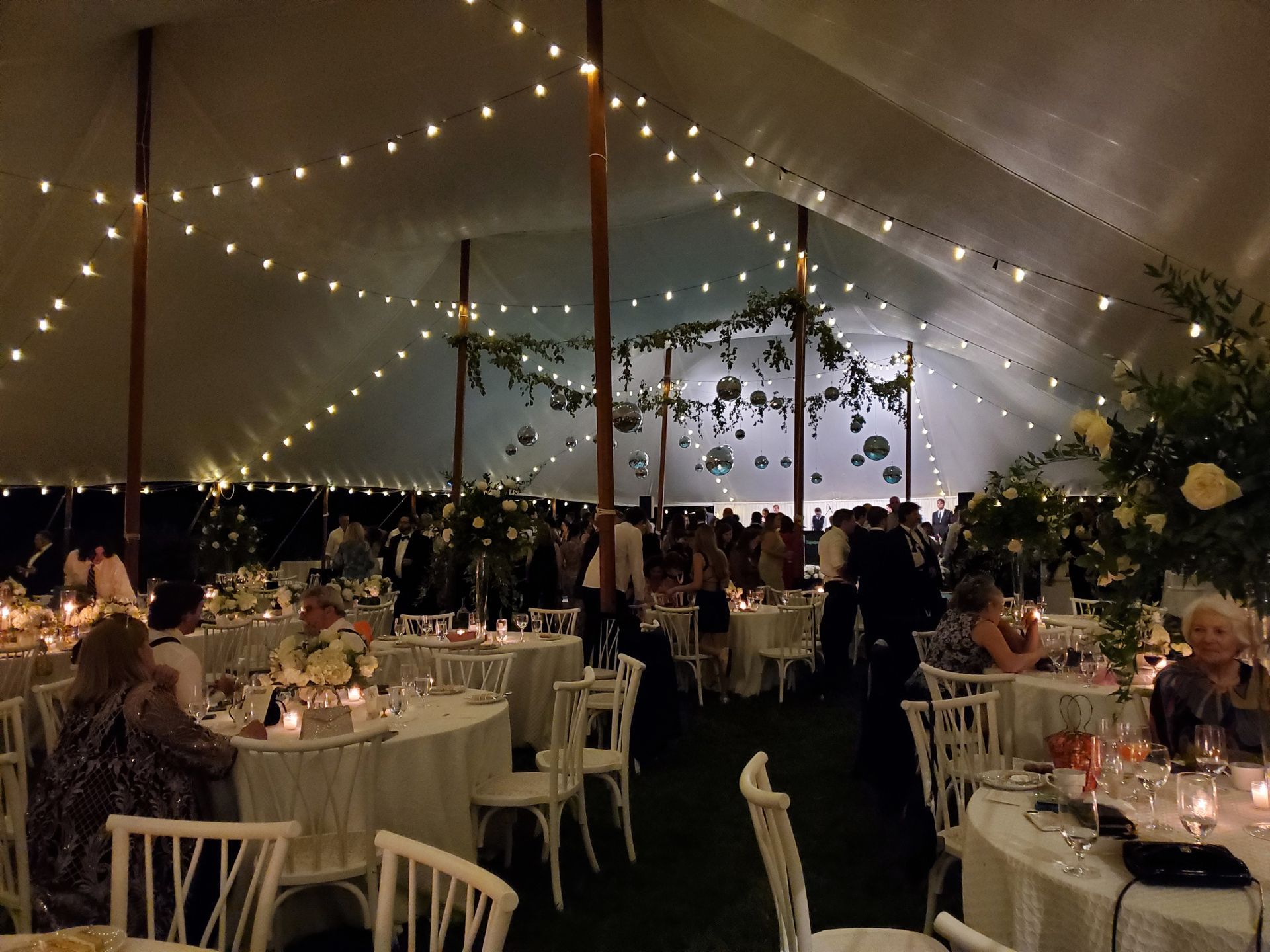 Wedding reception inside a tent with string lights, round tables, floral centerpieces, and guests.