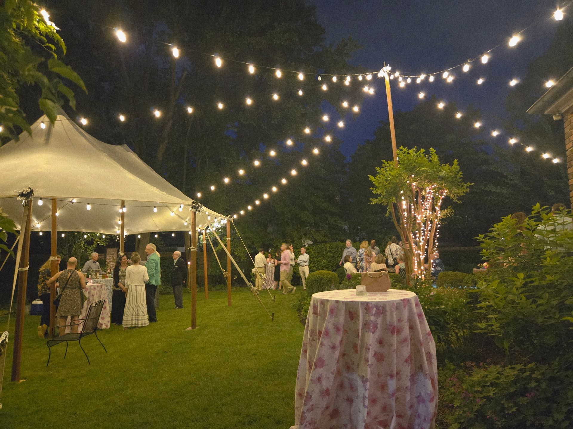 Outdoor evening wedding reception with string lights, tent, guests, and a table.