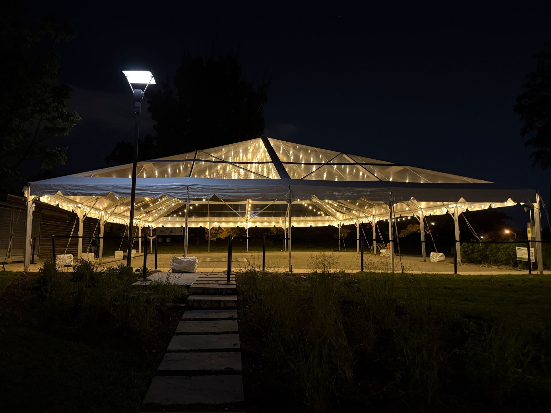 A lit-up clear tent at night with string lights, surrounded by grass and a pathway.