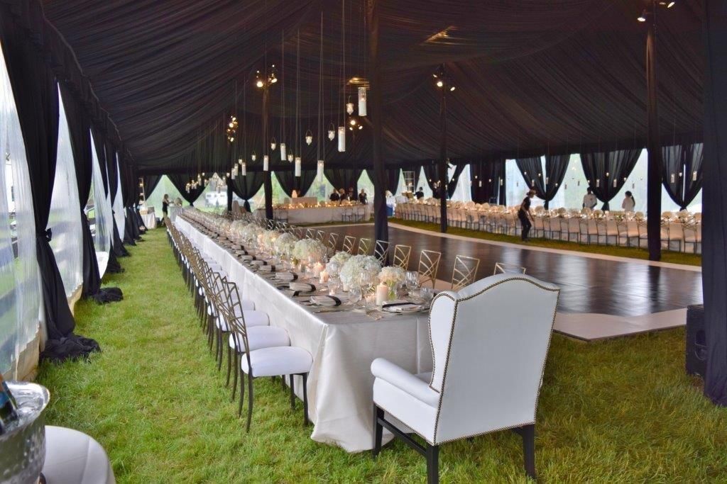 Long, elegant dinner table in a black tent, set on grass. White linens, chairs, and flowers.
