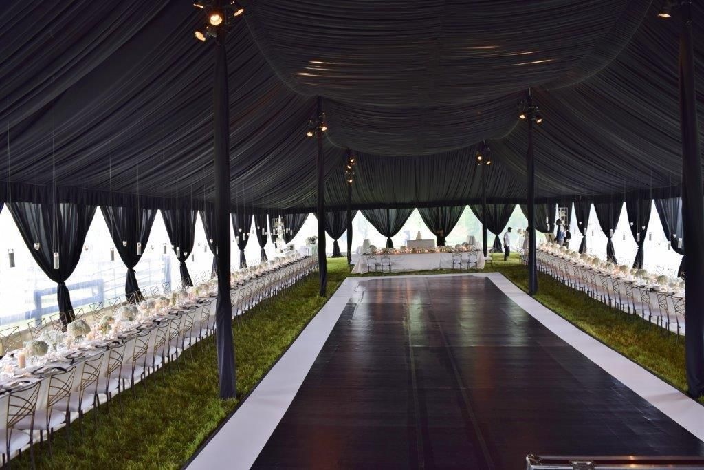Black-draped event tent with long tables set for a formal gathering. Green ground cover and a dark floor.