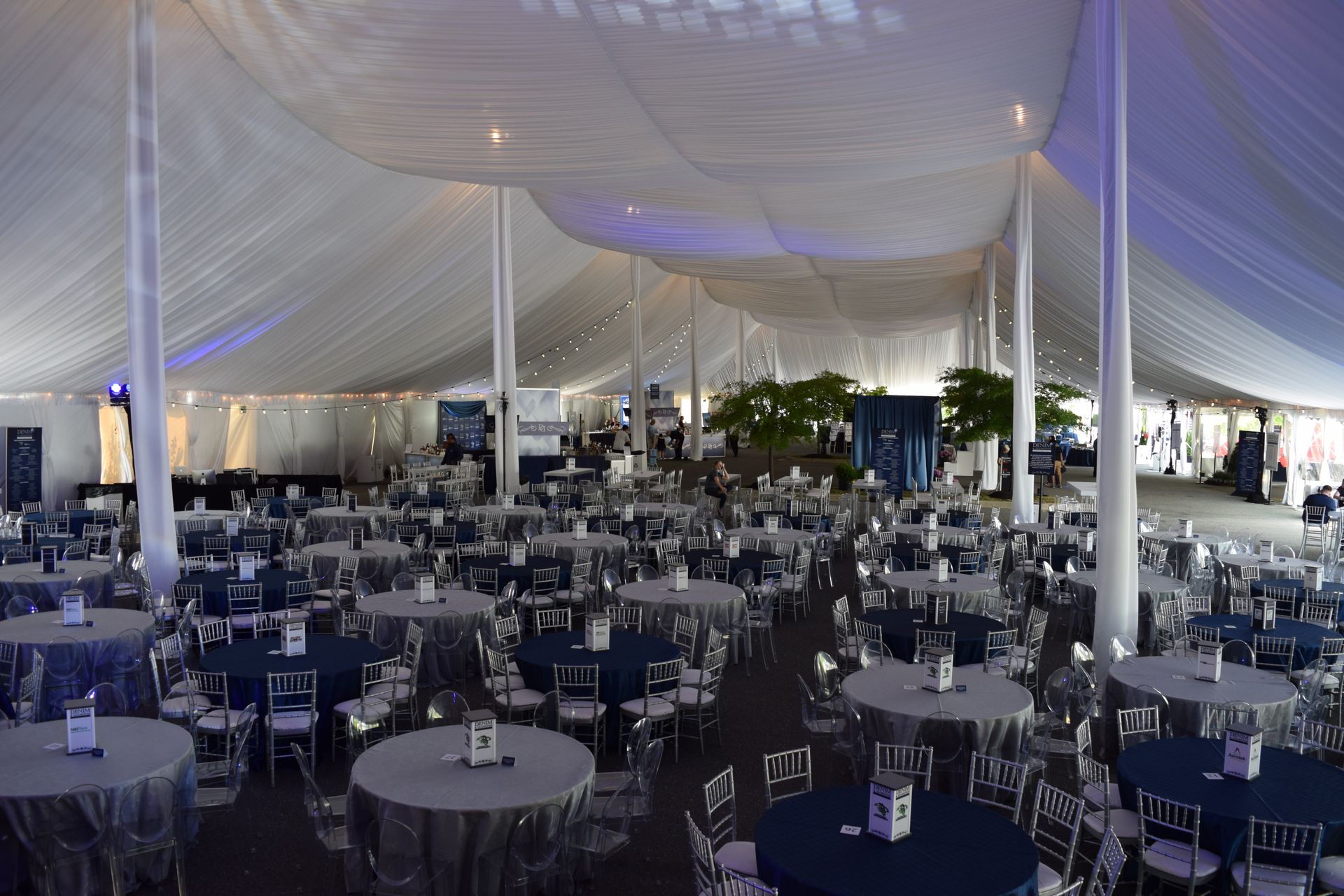 Large tent interior with rows of round tables, chairs, and blue tablecloths.