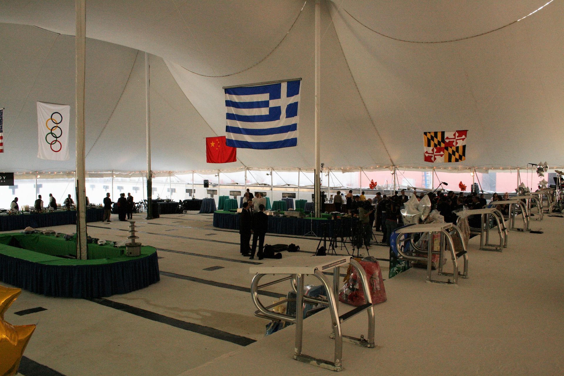 Inside a large white tent, people are seated at tables, flags include Greece, China, Maryland, and Olympic rings.