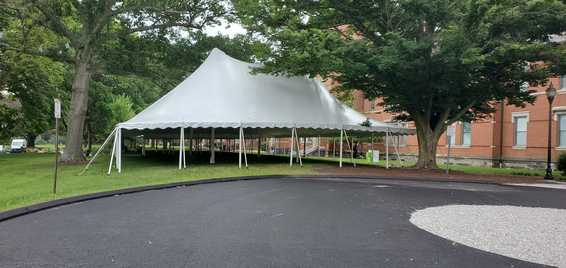 A large white tent set up on a grassy area next to a brick building, with trees and a paved road.