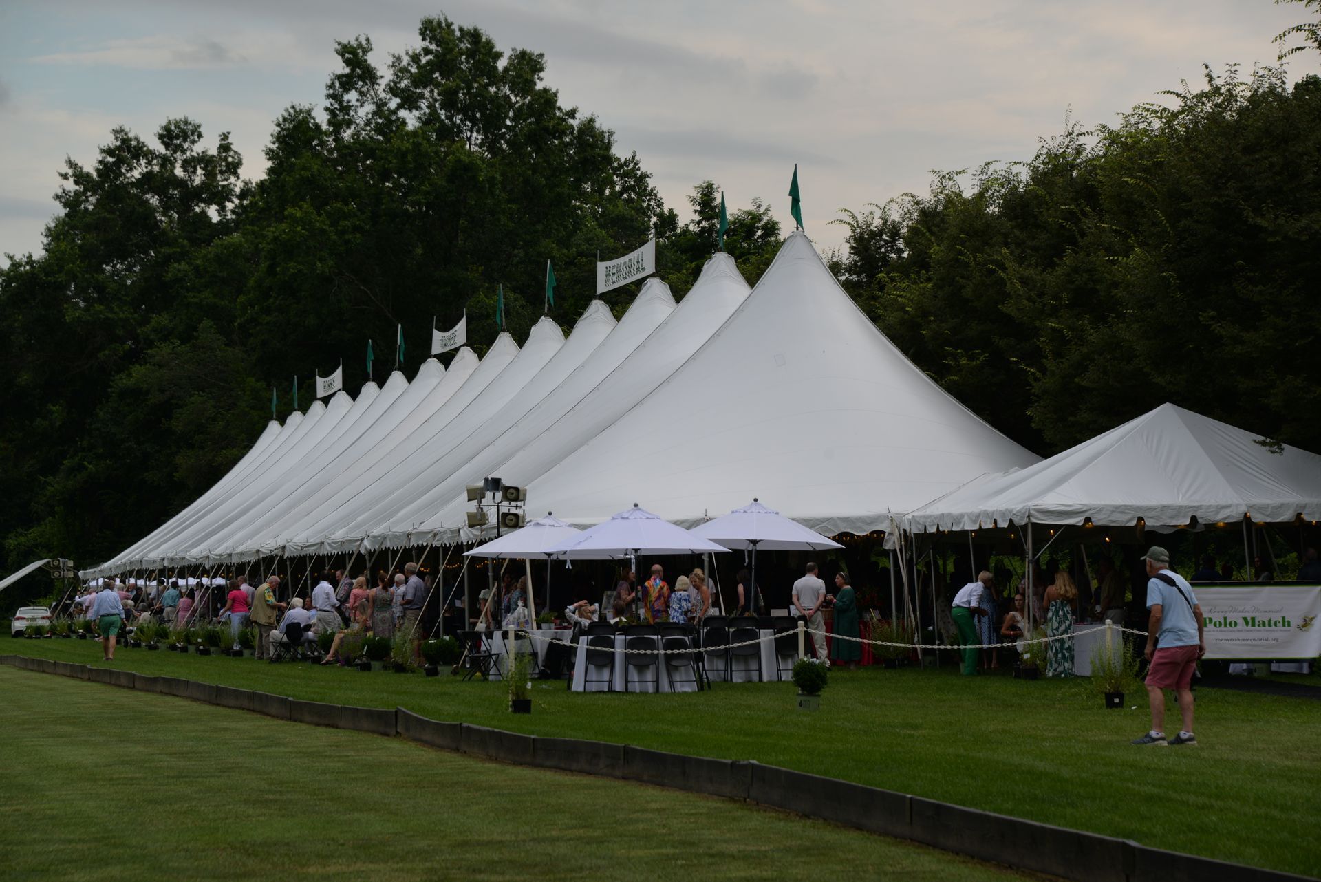 Large white tents with people on the lawn, trees in the background, event in progress.