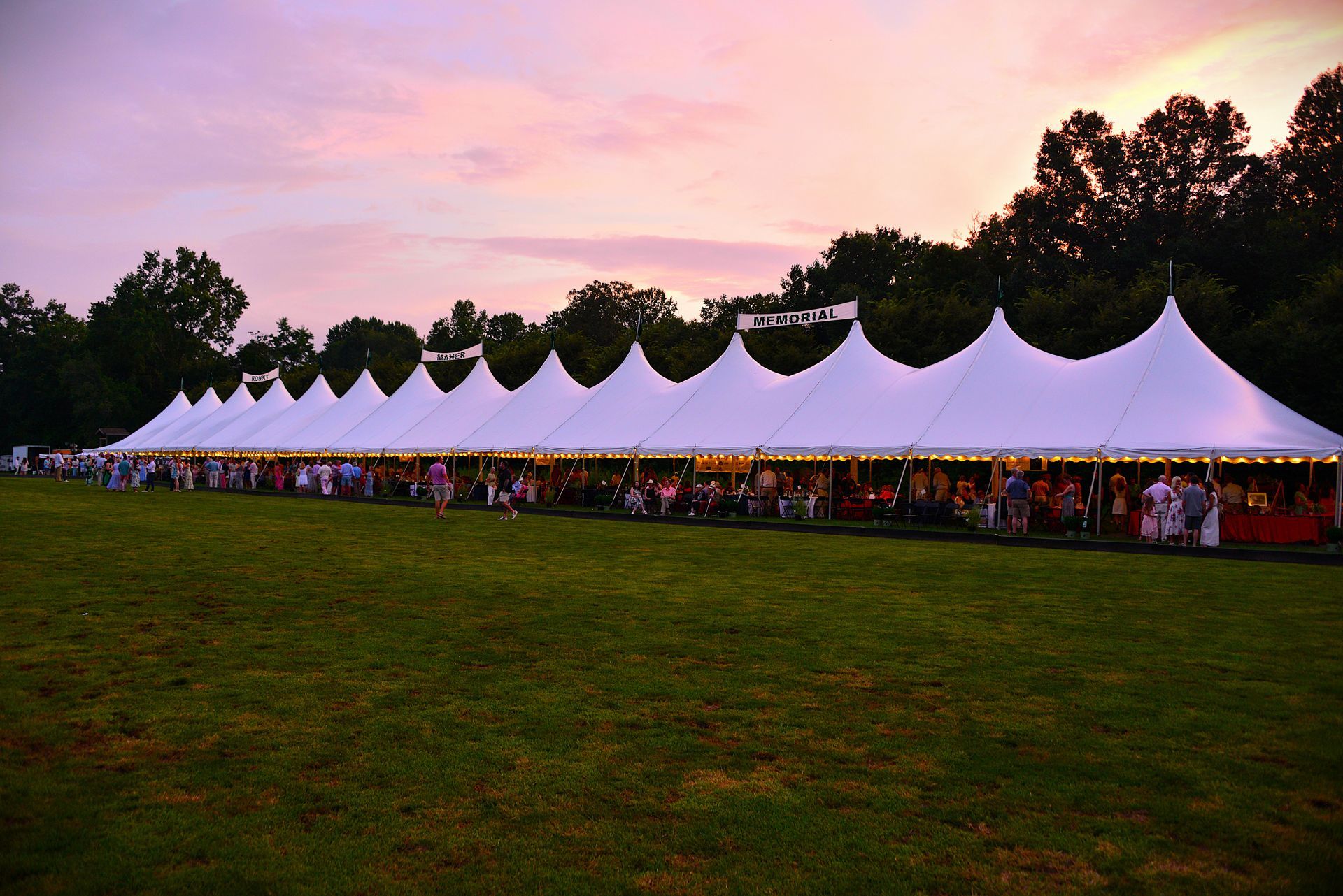 Long white tent at dusk, people milling inside, green grass, pink and purple sky.