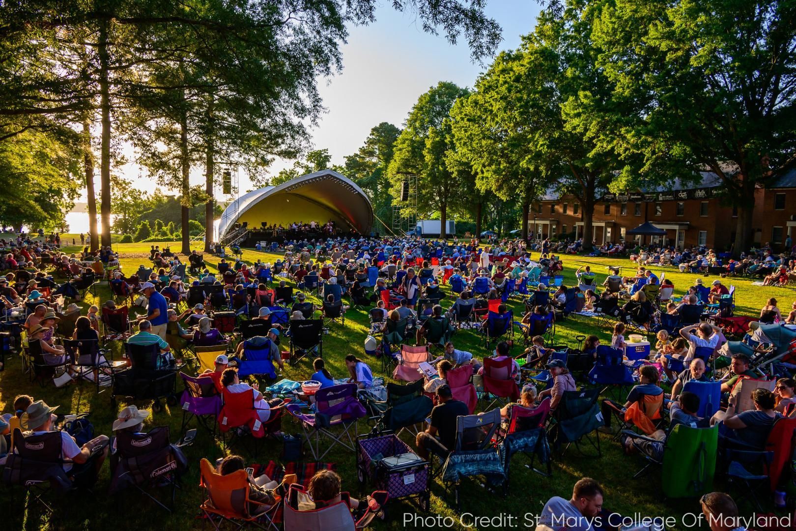 Large outdoor concert; crowd in lawn chairs faces stage. Trees, setting sun.