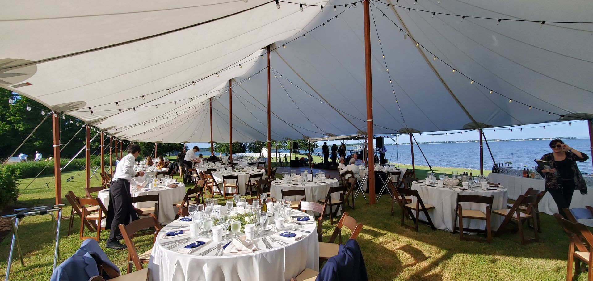 Wedding reception under a white tent on a grassy area near the water. Guests at tables.