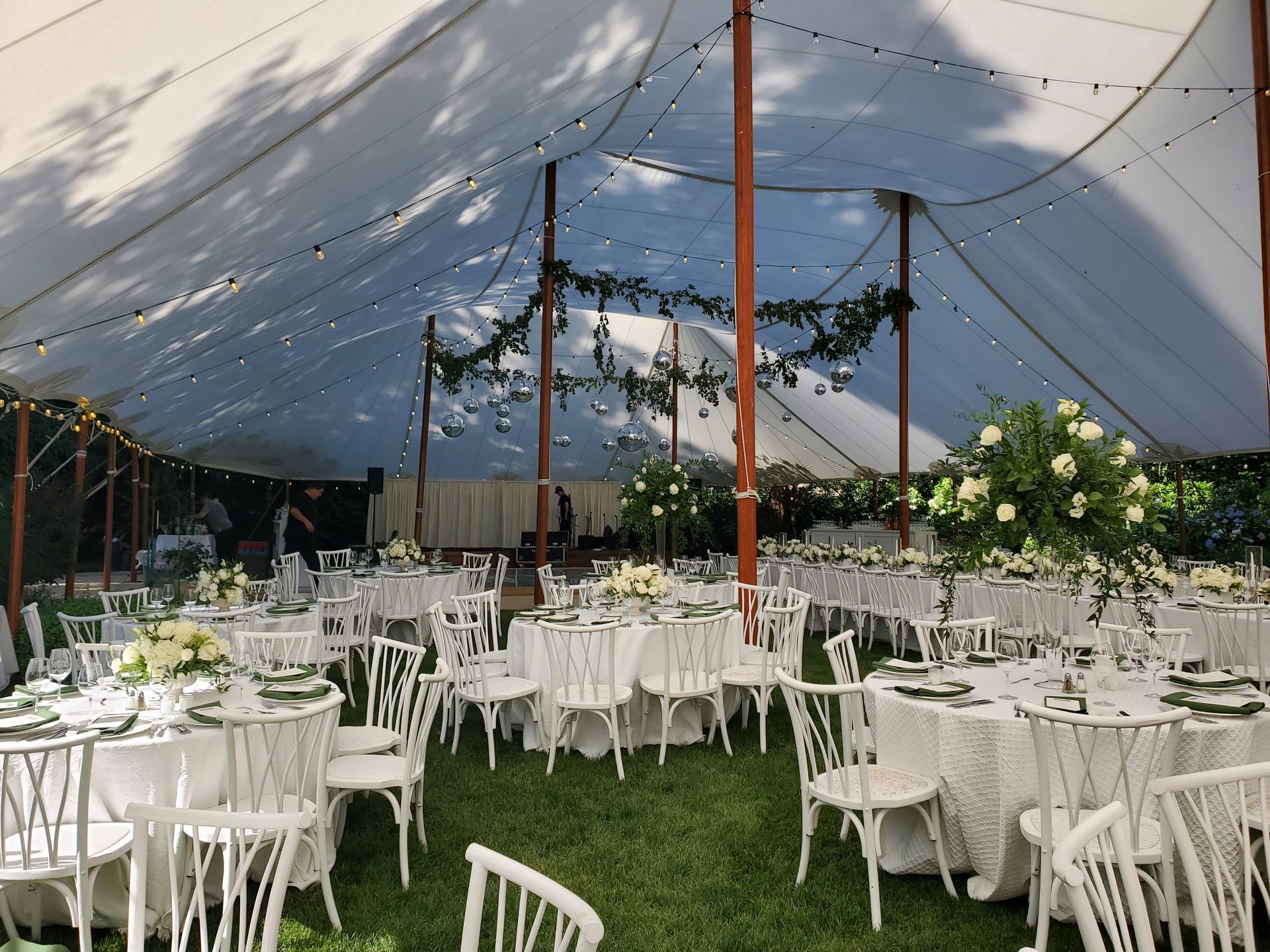 Wedding reception under a white tent. Tables set with white linens and floral arrangements. Greenery and string lights.