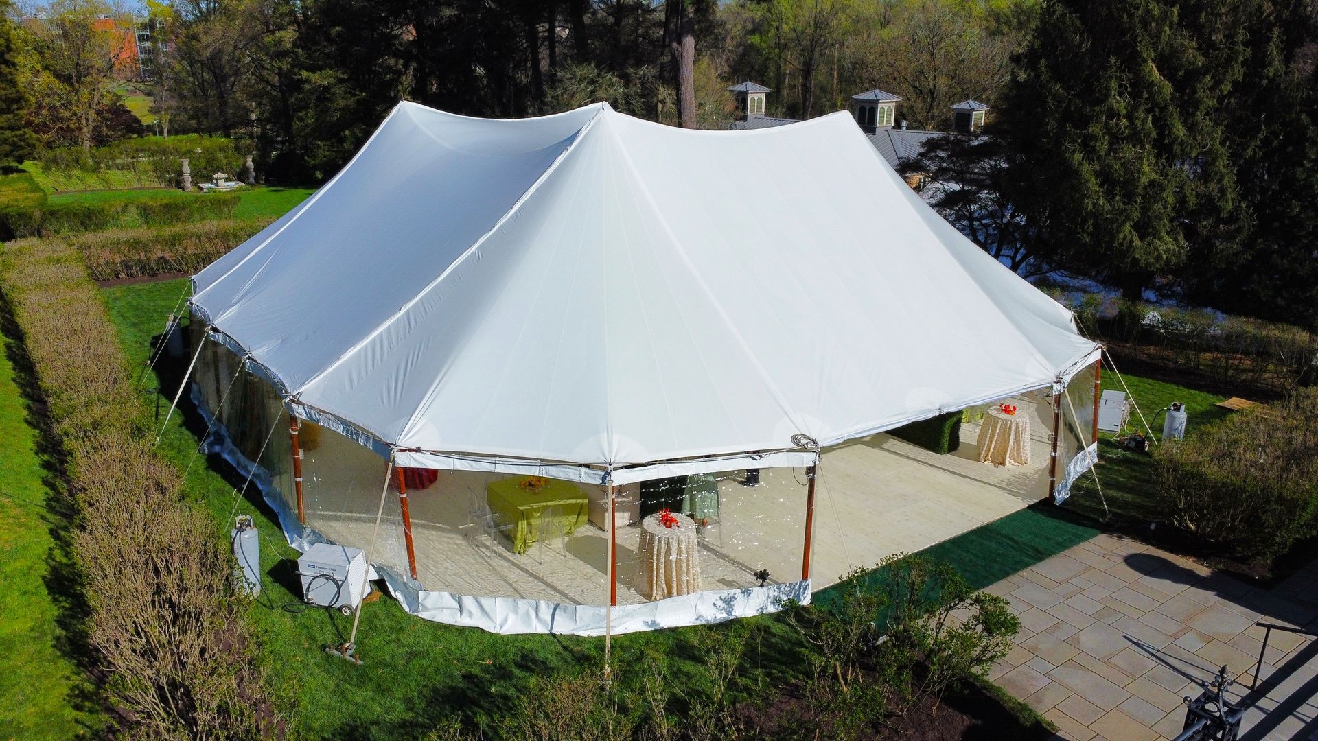 White tent set up on a grassy lawn, surrounded by trees. Clear side walls and wood supports visible.