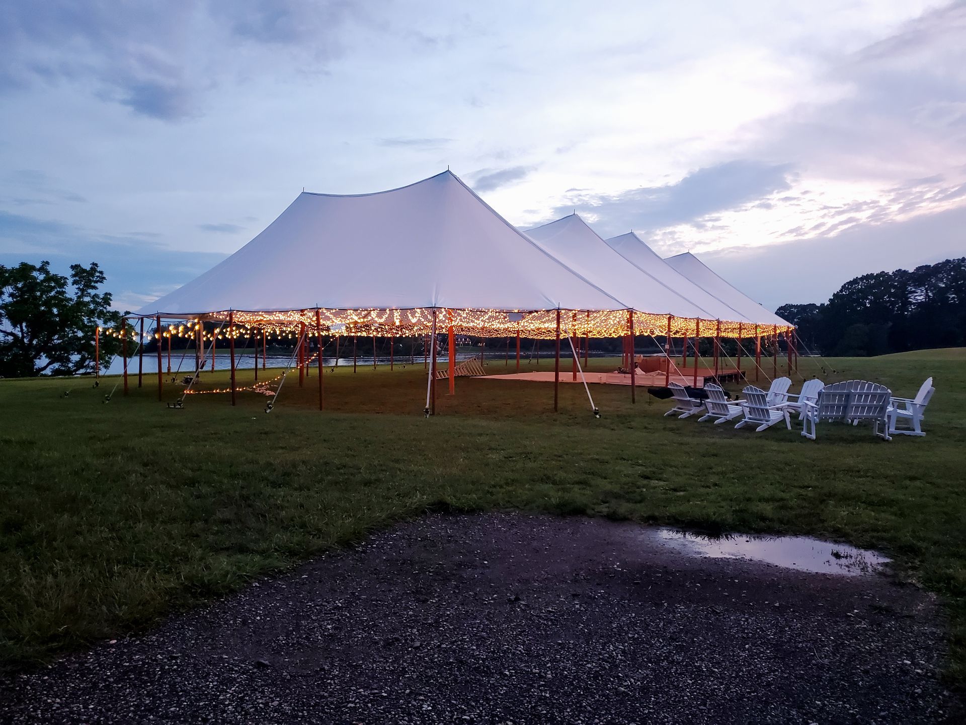 White tent lit with string lights, set on grassy field with tables and chairs. Dusk sky.