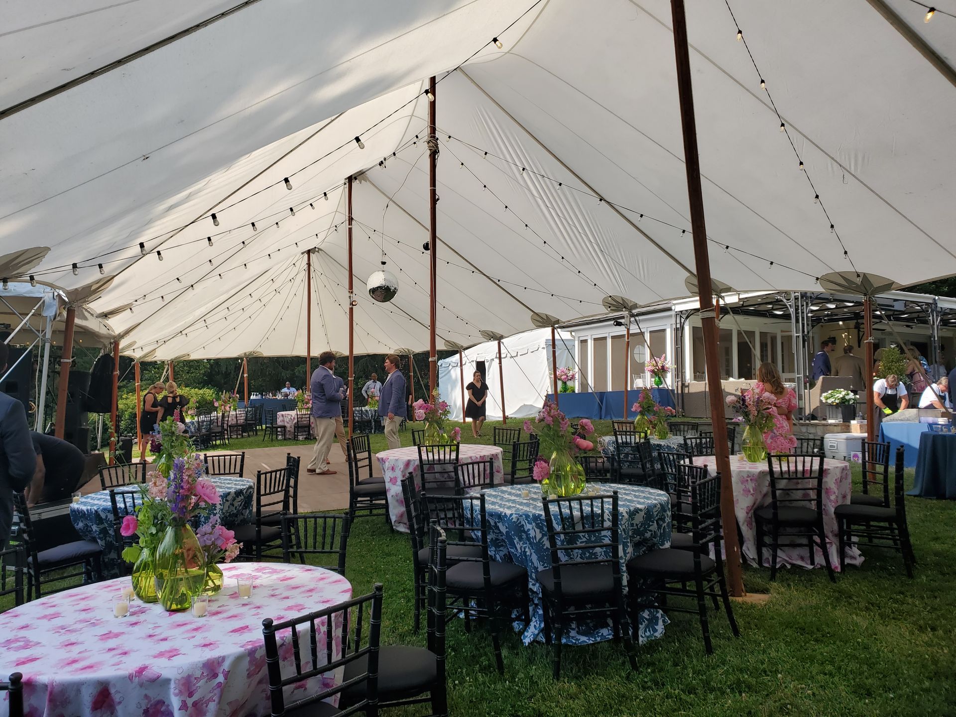 Outdoor tented event with round tables, floral tablecloths, and string lights. People are visible.