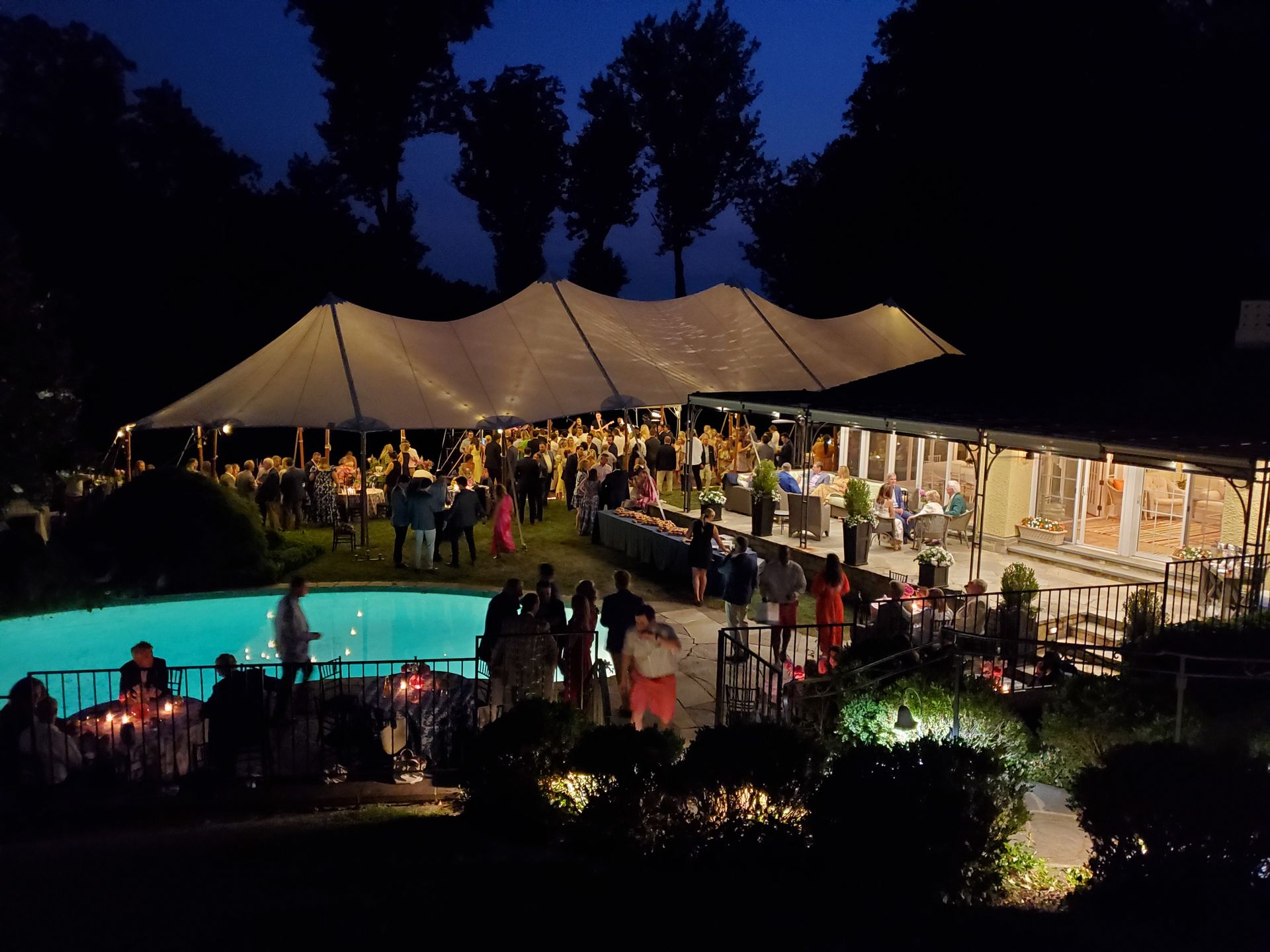 Nighttime outdoor event with tent, pool, and many people mingling.