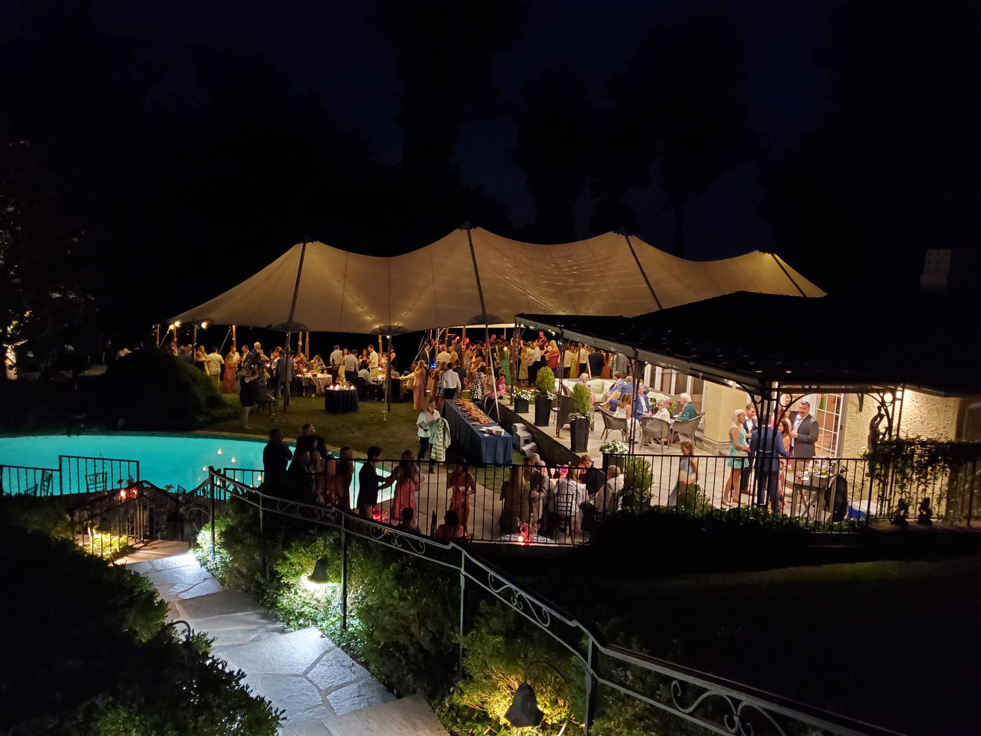 Nighttime outdoor event with guests under tent and near pool.