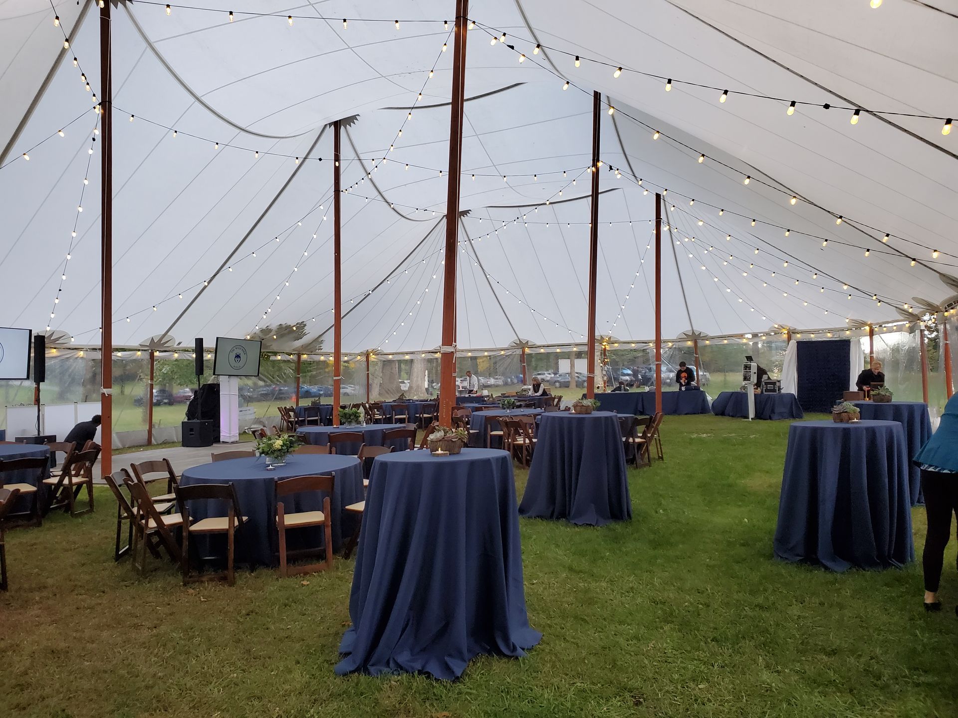 Inside a white tent, tables with navy blue cloths and chairs are set for an event on a grassy lawn.