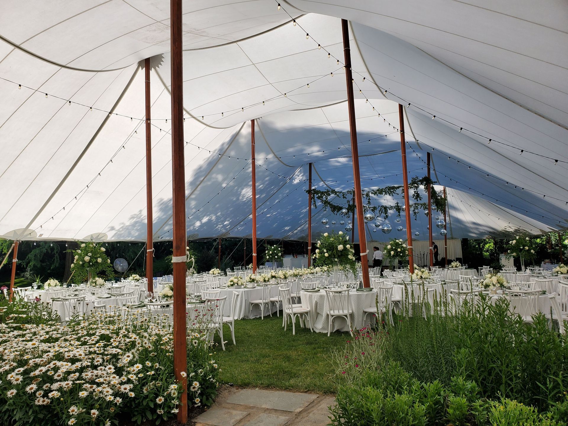 Wedding reception under a white tent. Tables and chairs are set up on grass, with flowers and wooden poles.