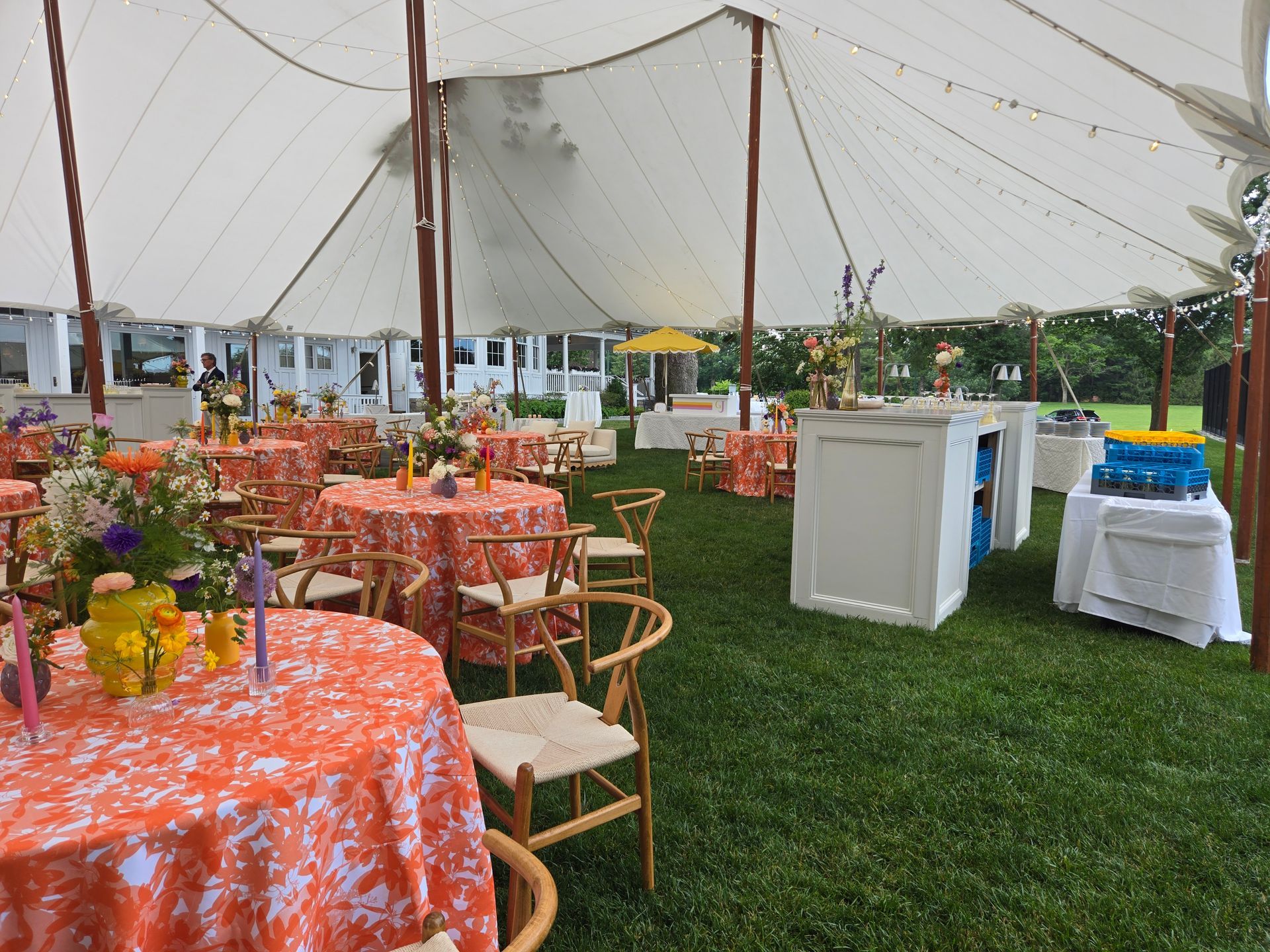 Outdoor party under a white tent with orange tablecloths, wooden chairs, and floral centerpieces.