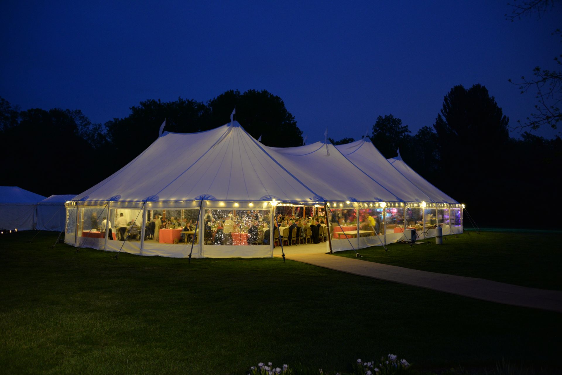 Large white tent lit up at night on a lawn, with people visible inside.