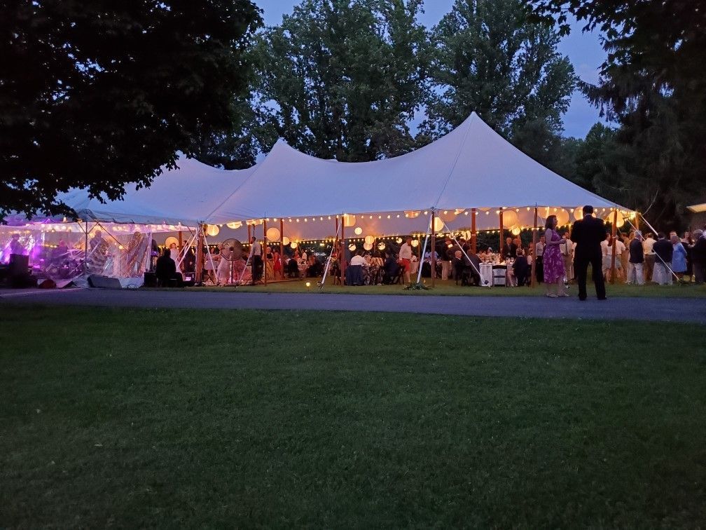 Wedding reception under a large white tent at dusk, with guests, lights, and a green lawn.