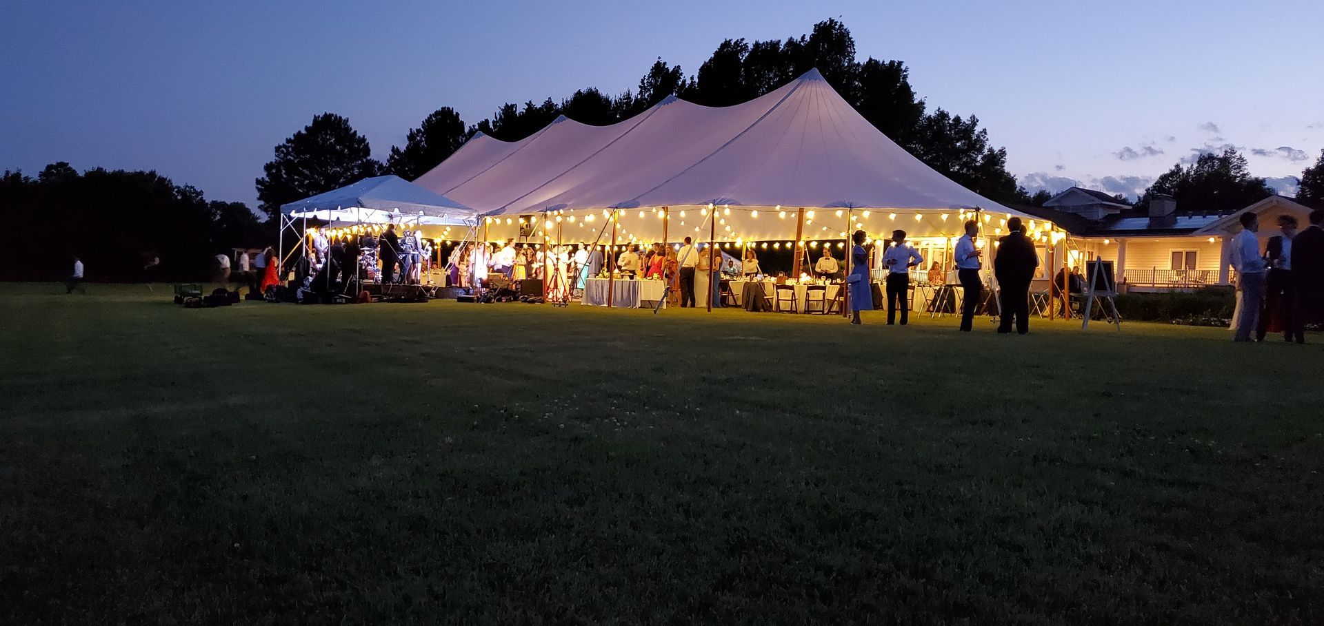 A large white tent at an outdoor event at dusk, with people gathered and lights strung.