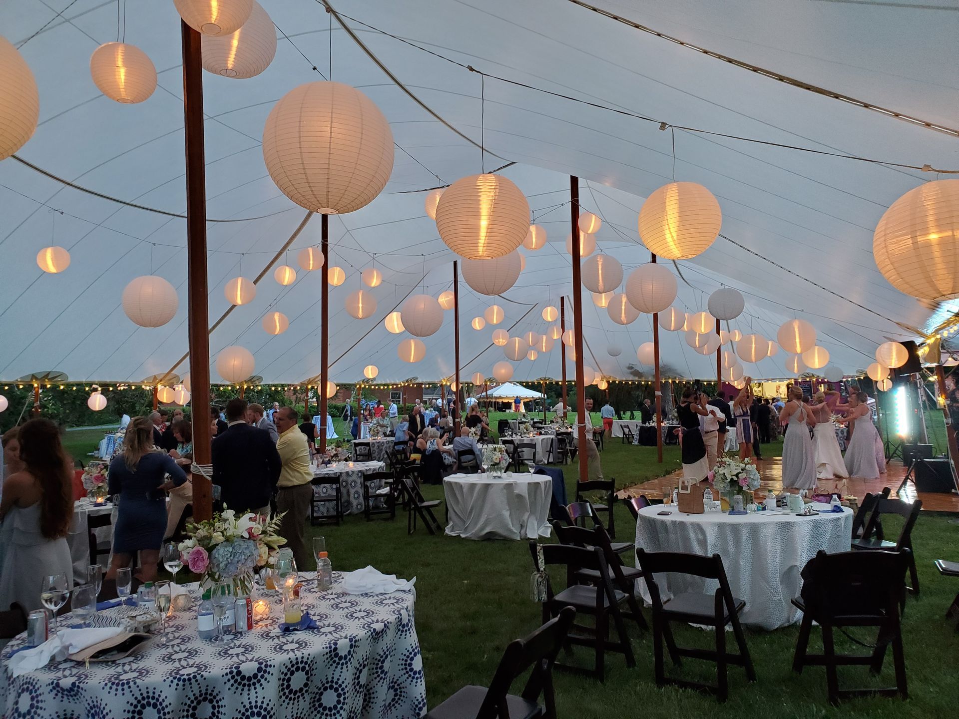 A large tent with round paper lanterns, tables with people, and a wedding reception in a grassy area.