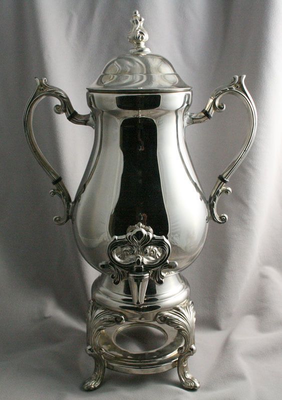 Silver coffee urn with ornate handles, lid, and base on a light background.