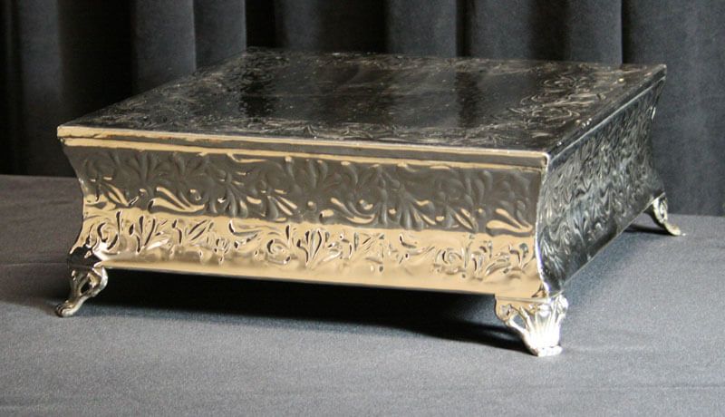 Silver, ornate cake stand with four claw feet, floral border, set on a gray surface.