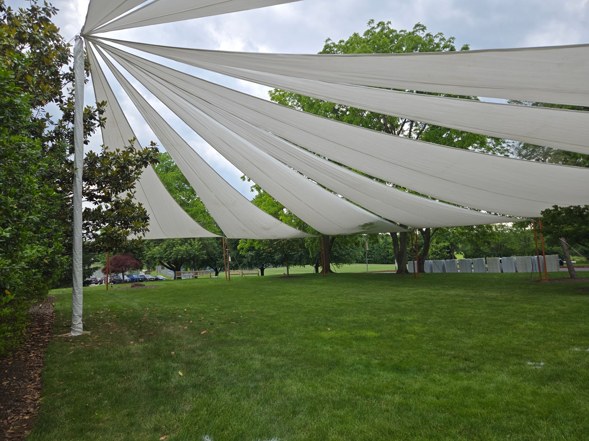 White shade sails over a green lawn, trees in the background, overcast sky.