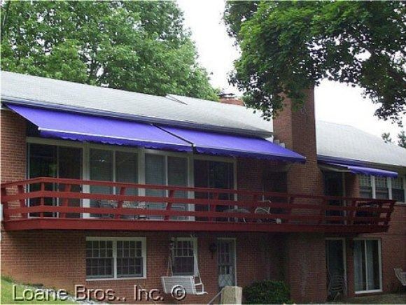 A two-story brick house with a red deck, blue awnings, and a tree.