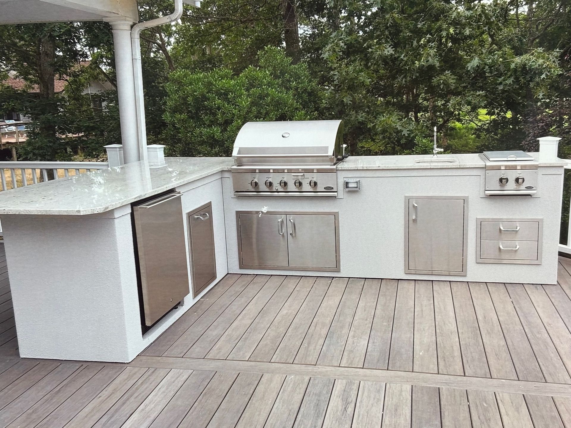 Outdoor kitchen with grill, refrigerator, and sink on a wooden deck. White countertop and cabinets.