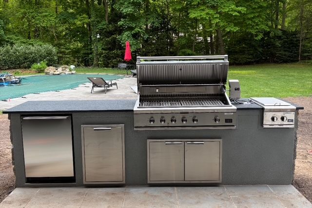 Outdoor kitchen with a grill, refrigerator, and storage cabinets in a backyard.