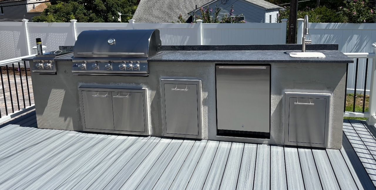 Outdoor kitchen on a deck with grill, sink, and storage cabinets. Gray concrete base with stainless steel appliances.