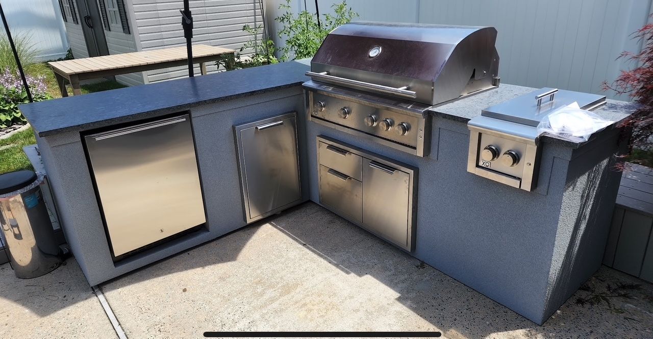 Outdoor kitchen with stainless steel grill, refrigerator, and sink on a gray concrete structure.