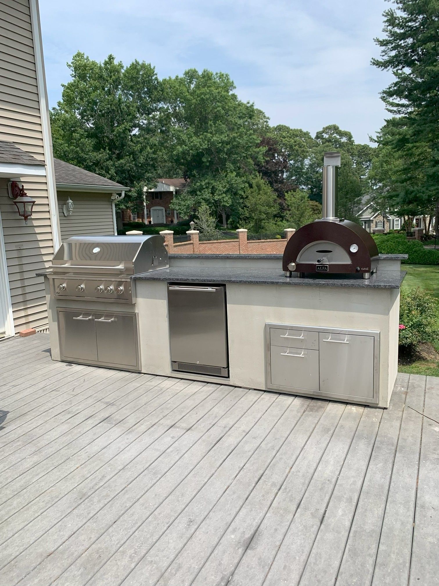 Outdoor kitchen with a grill, pizza oven, refrigerator, and storage on a wooden deck.