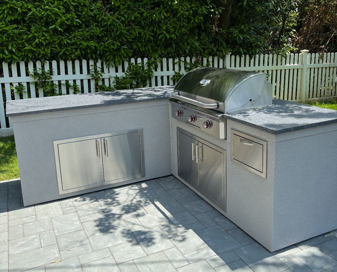 Outdoor kitchen with stainless steel grill, cabinets, and counter, on a paved patio with a white picket fence backdrop.