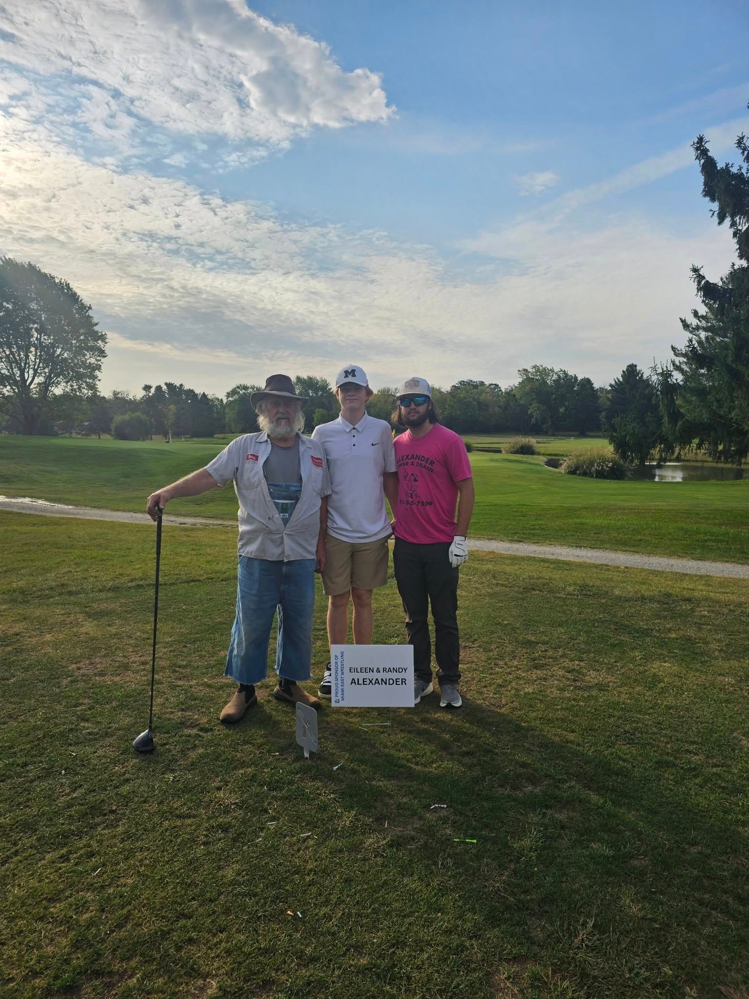 Three people posing on a golf course with clubs and a sign under a cloudy sky.