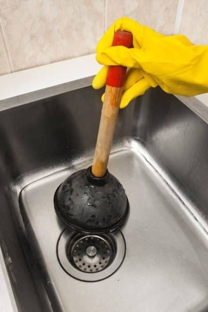 Gloved hand using a toilet plunger in a stainless steel sink