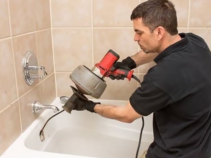 Plumber using a drain auger to clear a bathtub drain in a tiled bathroom