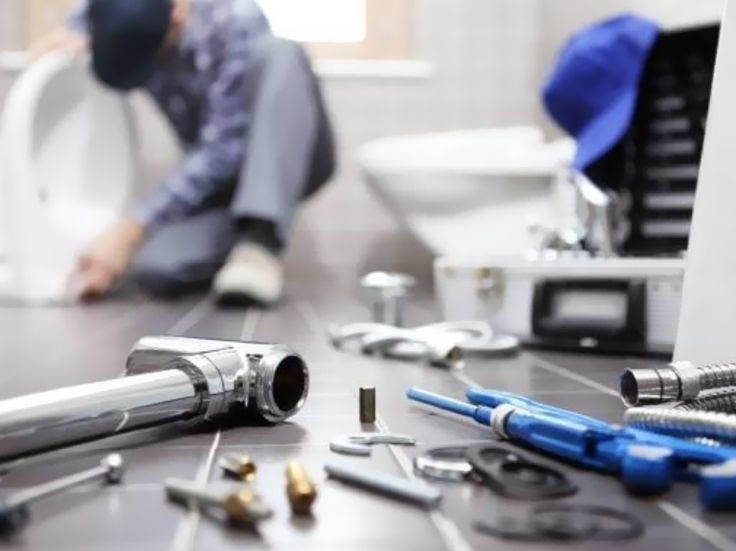 Scattered plumbing tools on a floor with a blurred worker repairing a sink in the background