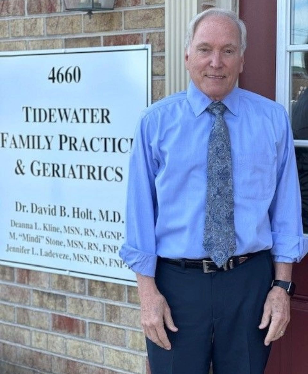 dr. david holt  in a blue shirt and tie standing in front of a sign for tidewater family practice and geriatrics