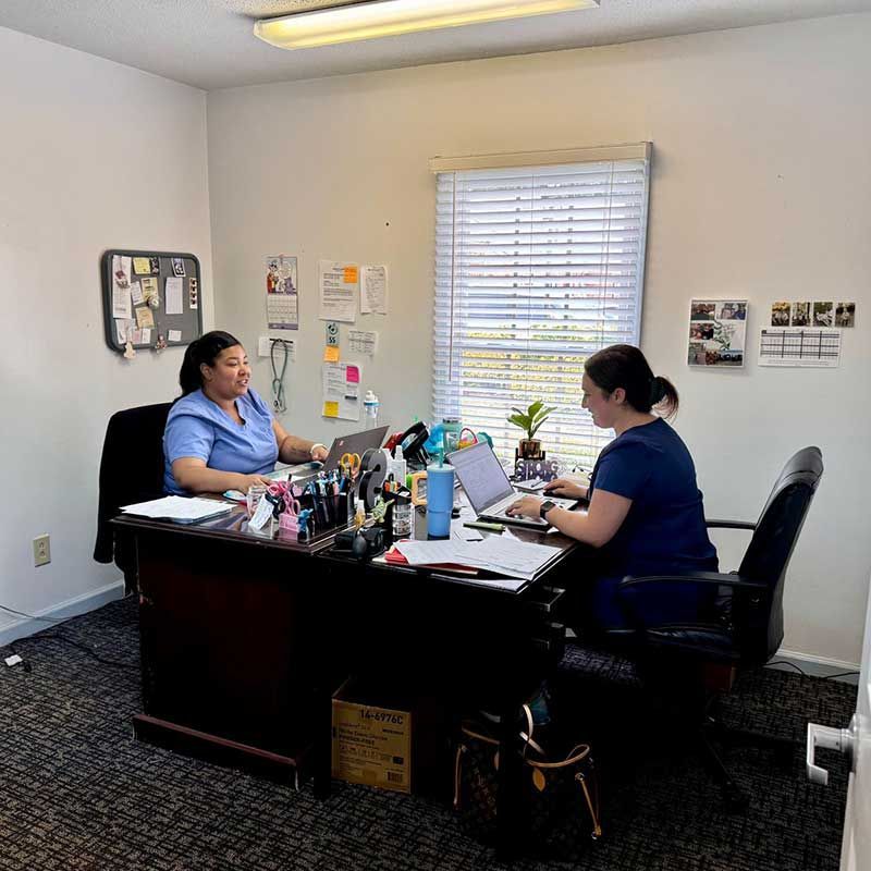 two women are sitting at a desk with laptops in an office